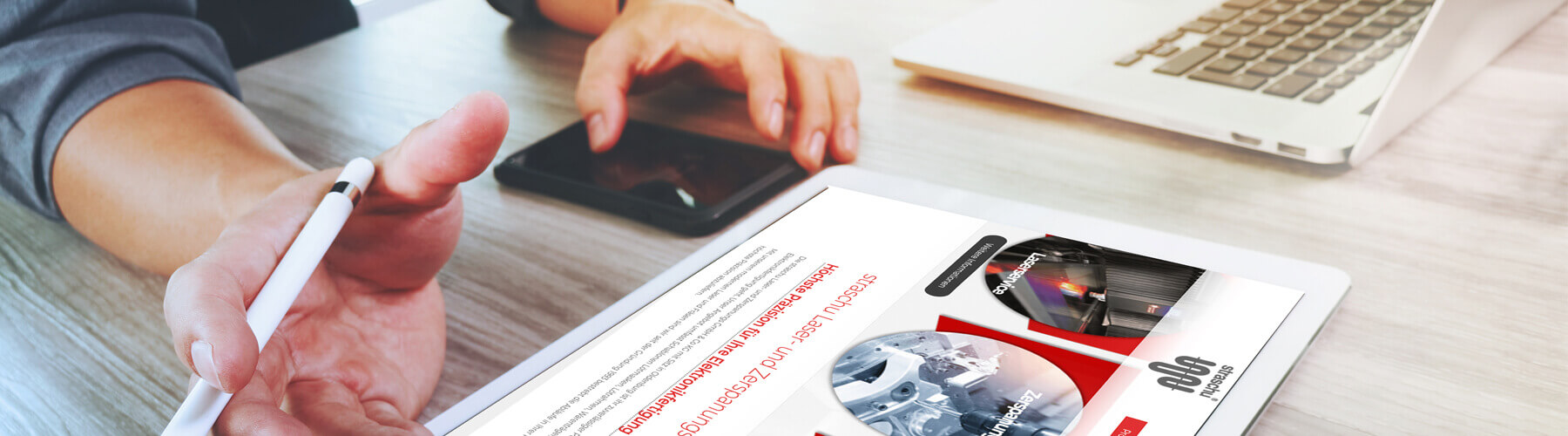 Auf einem Tisch liegen im Vordergrund ein Tablet mit der straschu Laser- und Zerspanungs-Website und ein Mann hat seine Hand mit einem Tablet-Stift darüber. Mit der anderen Hand tippt er auf einem Handy, welches hinter dem Tablet liegt und im Hintergrund ist ein aufgeklappter Laptop zu sehen.