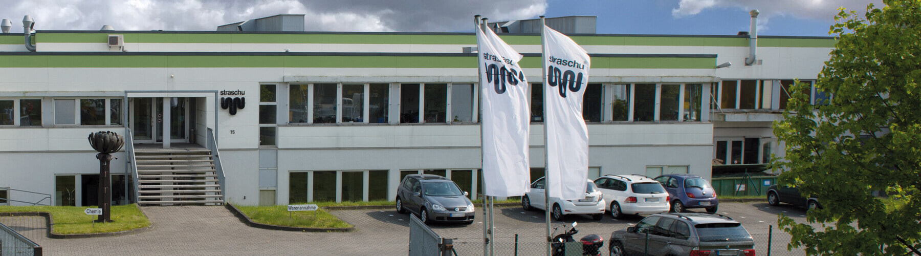 Ein weißes Gebäude mit Flachdach und einem Parkplatz davor, an der Auffahrt wehen zwei straschu-Fahnen im Wind.